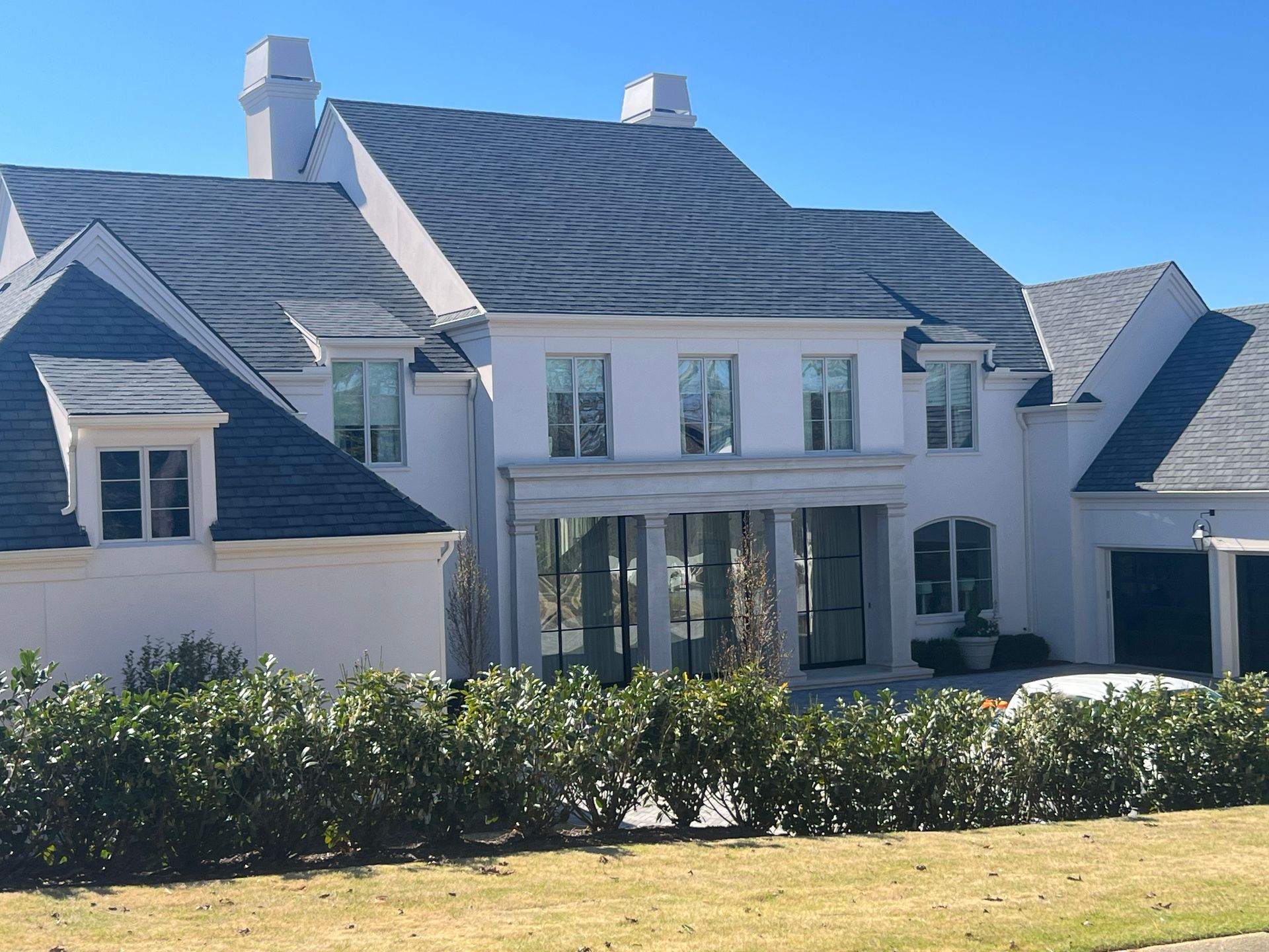 White two-story house with dark gray roof, columns, windows, and manicured hedge in front. Sunny day.