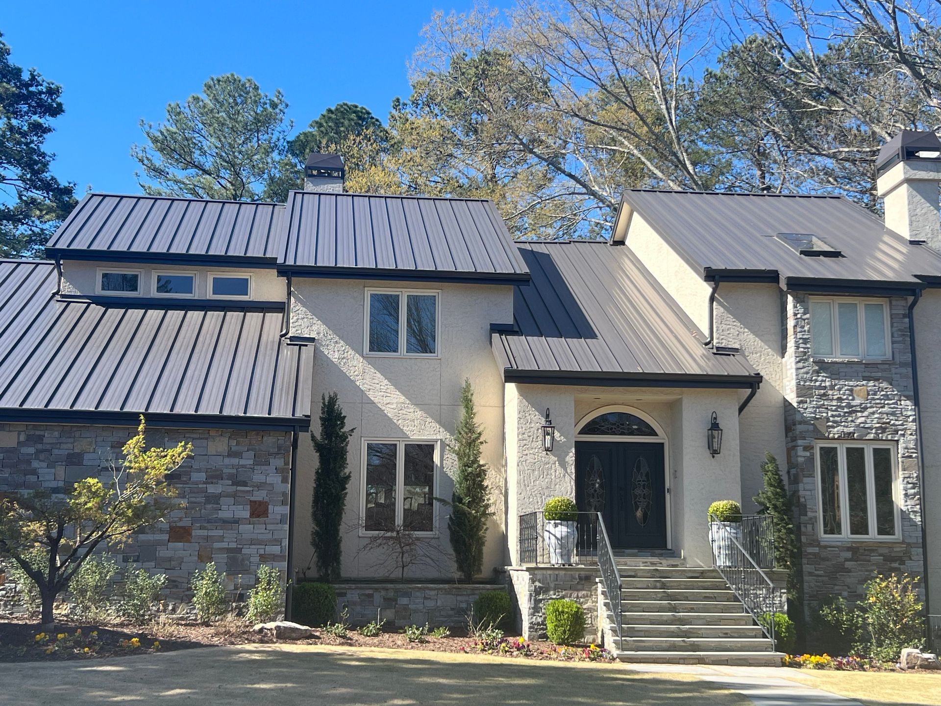 Modern two-story house with a dark metal roof, stone and stucco facade, and manicured landscaping.
