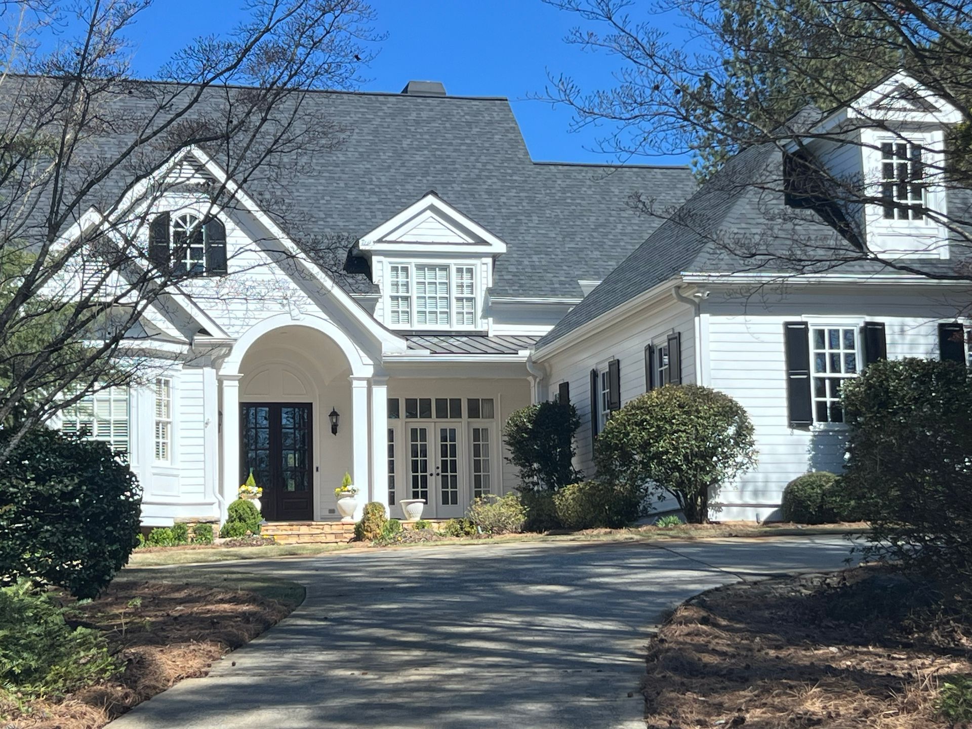 White house with black shutters, dormers, and dark gray roof; a driveway leads to the front door.