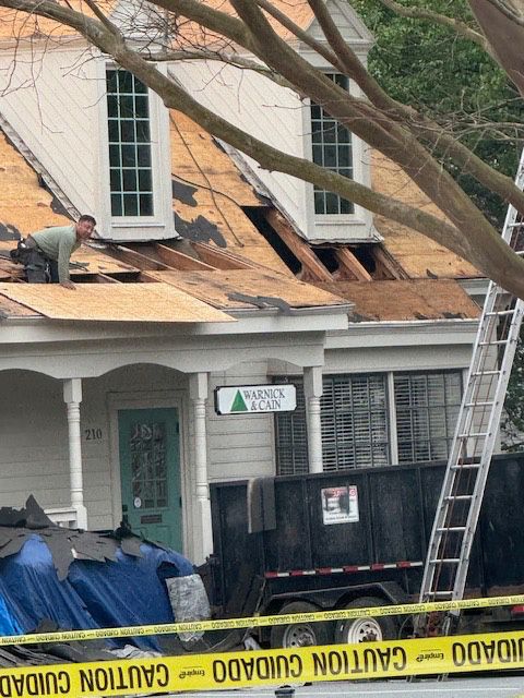 Man on roof removing shingles. House with open roof, ladder, and dumpster below. Caution tape present.