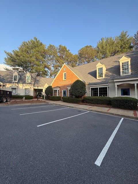 Parking lot in front of a brick building with a pitched roof, trees, and clear sky.