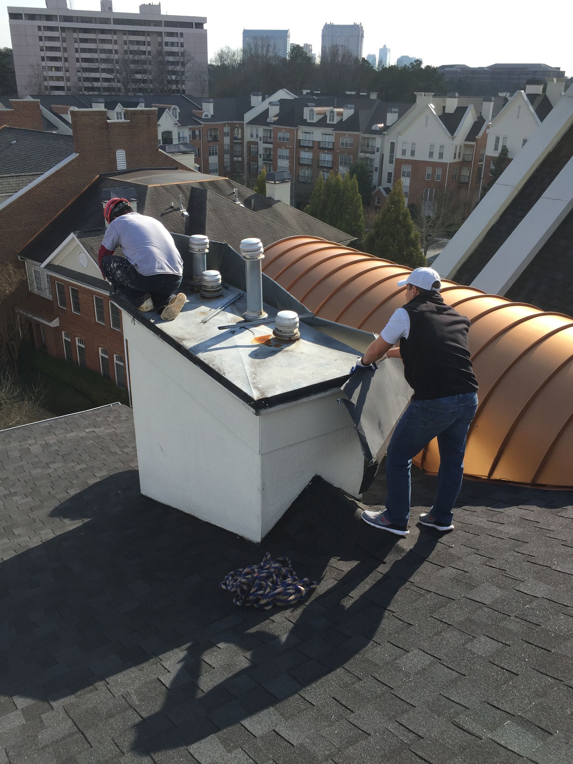 Two workers on a rooftop, one adjusting roofing material near a chimney, sunny day.