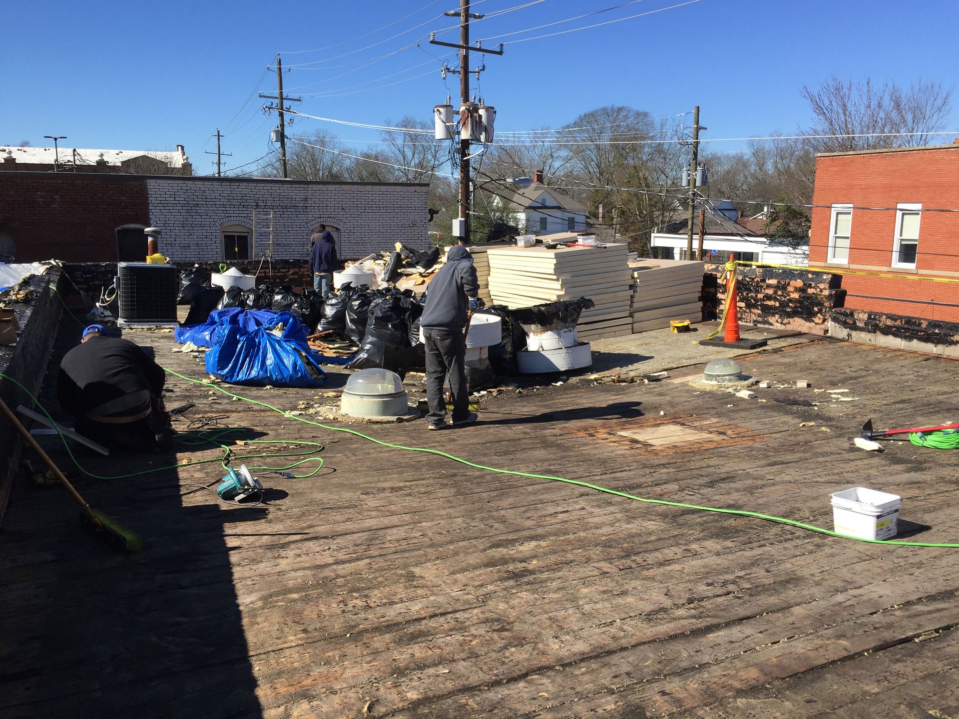 Construction workers on a flat roof with materials and debris. Blue tarp, black bags, and beige insulation are visible.