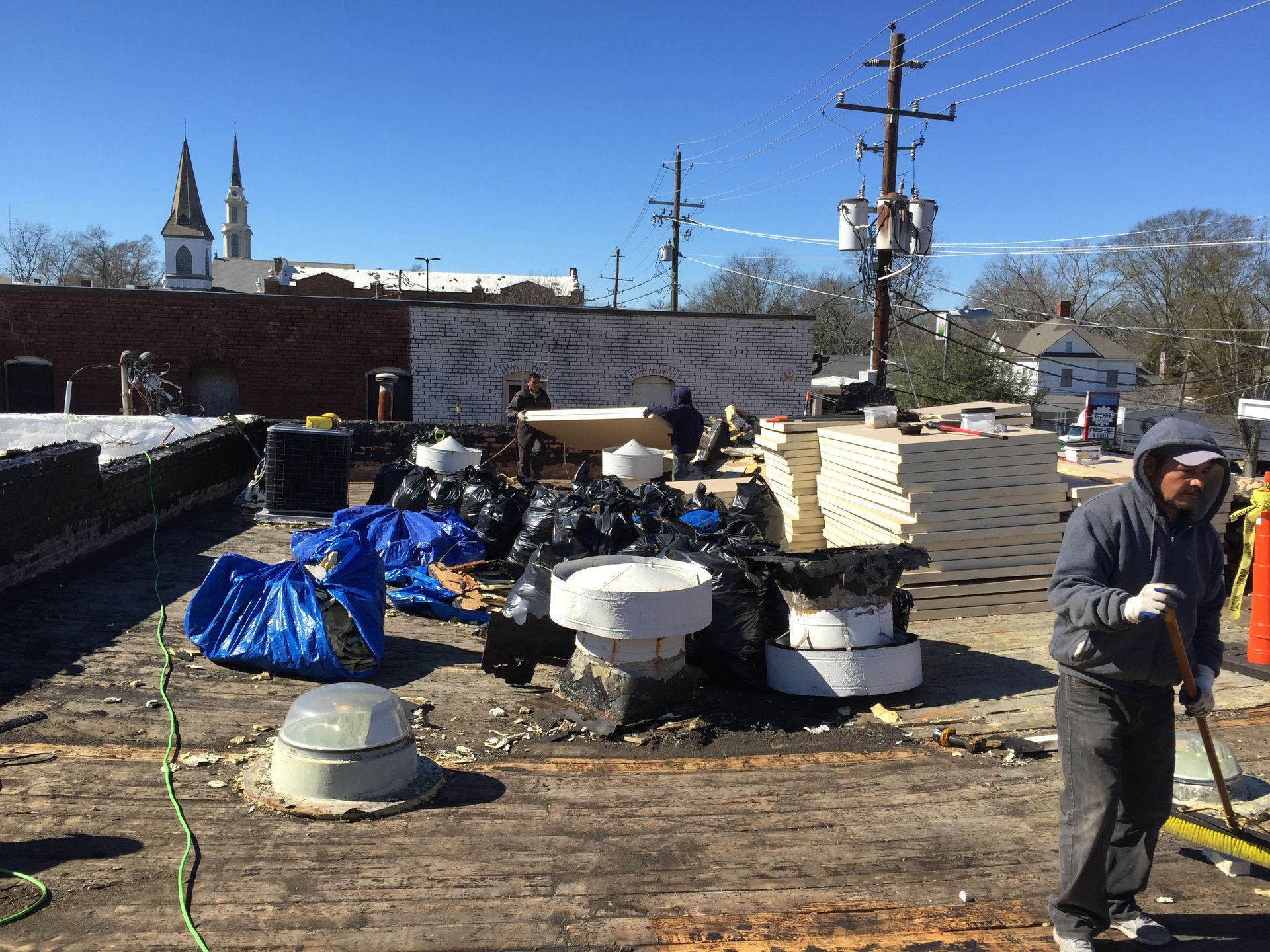 Roof repair site with workers, debris, materials, and a church in the background on a sunny day.