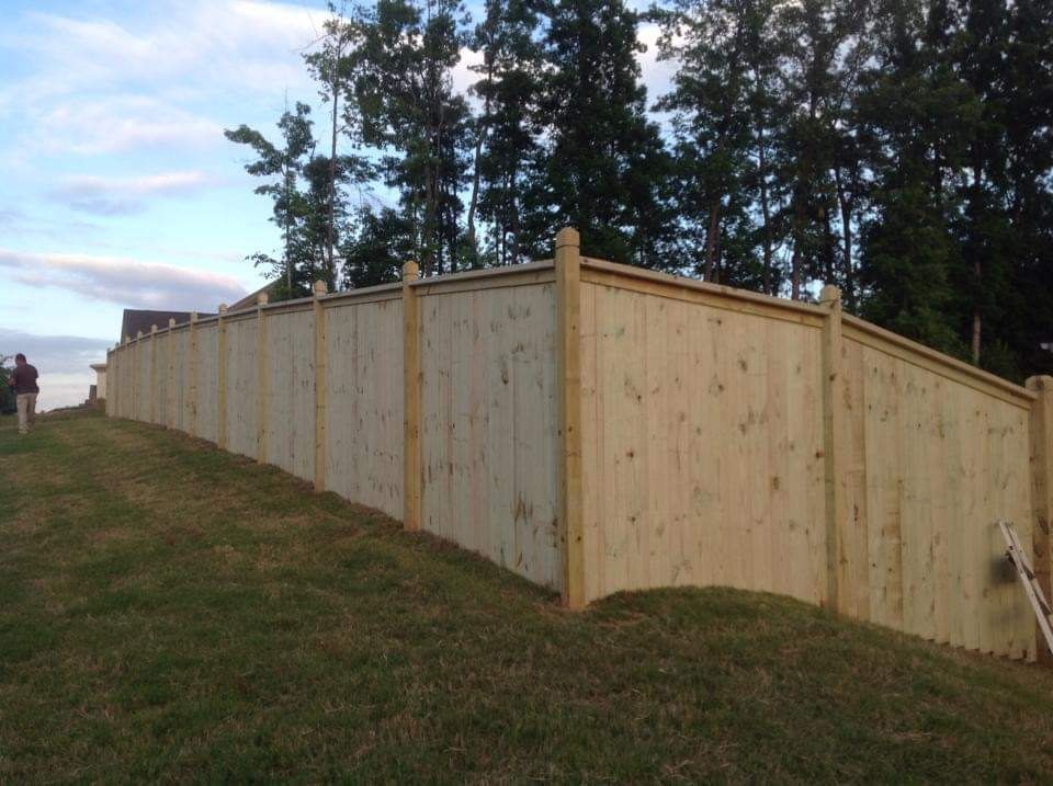 Wooden privacy fence along a grassy slope, with tall trees and a cloudy sky behind it