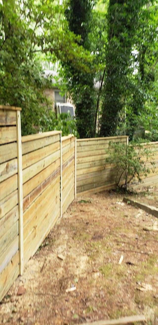 Wooden fence panels lining a narrow dirt yard with trees and shrubs in the background