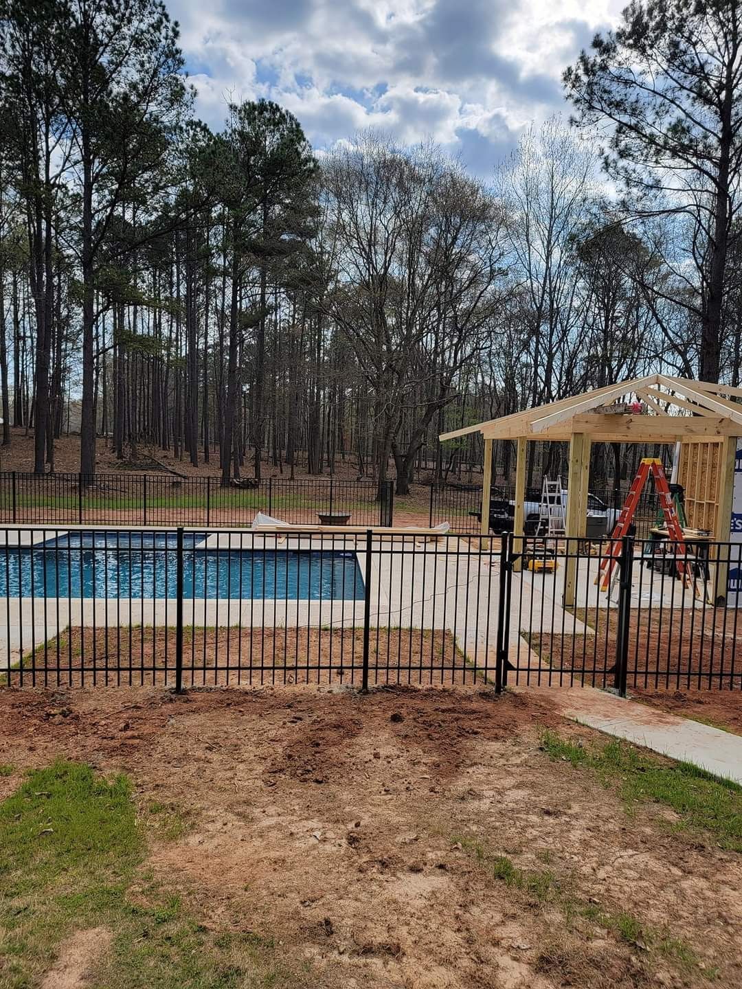 Backyard pool behind a black fence, with a pavilion and trees under a cloudy sky.