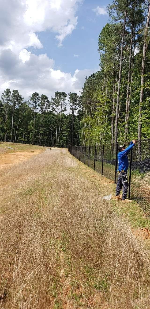 Person spraying a fence along a grassy field beside a wooded area under a blue sky