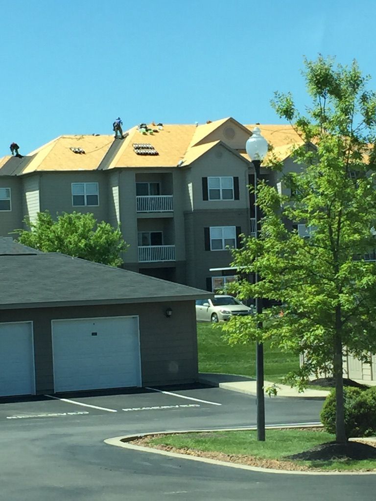 Apartment building with roofers on the roof under a blue sky, and a tree in front.