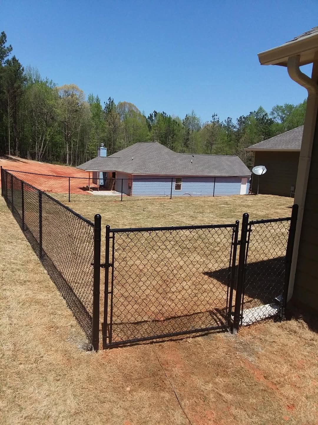 Chain-link dog pen beside a house in a fenced backyard, with a pool and woods in the background
