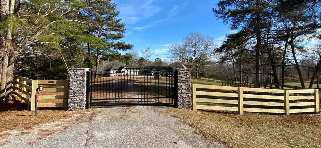 Closed black gate at the end of a gravel driveway, flanked by wooden fence posts and trees