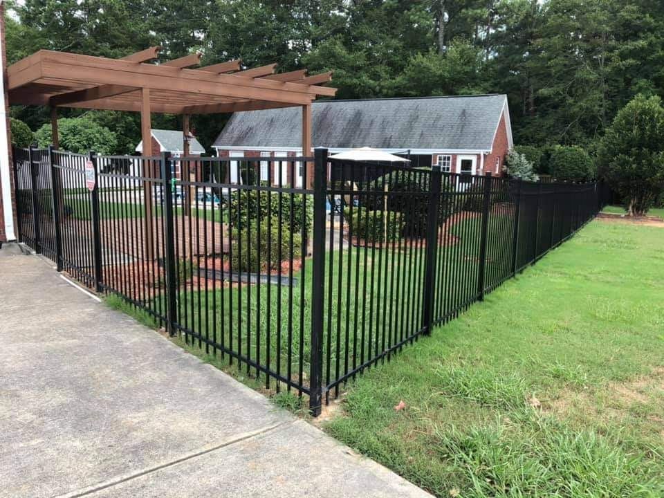 Black metal fence enclosing a backyard patio with pergola, garden beds, and a small shed beside a driveway.