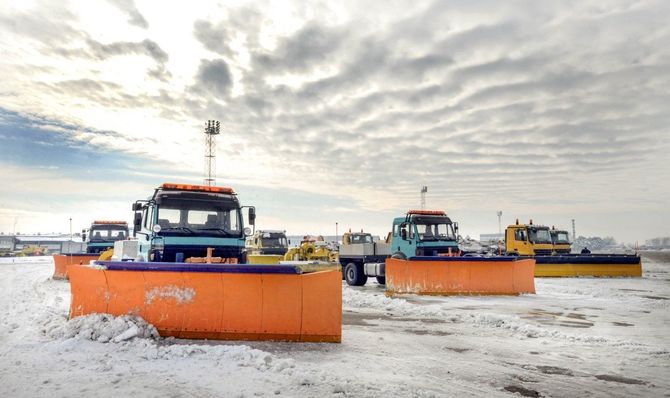 Three orange snowplow trucks lined up on a snowy field under a cloudy sky
