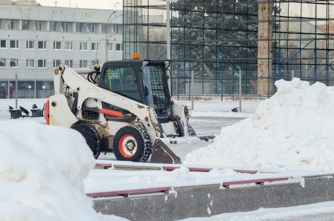 Skid steer clearing snow in a parking lot beside a building and large snow piles
