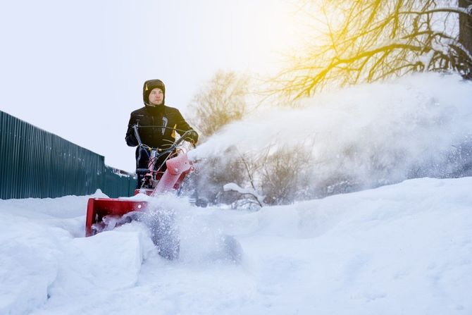 Person driving a red snowblower through deep snow beside a fence on a sunny winter day
