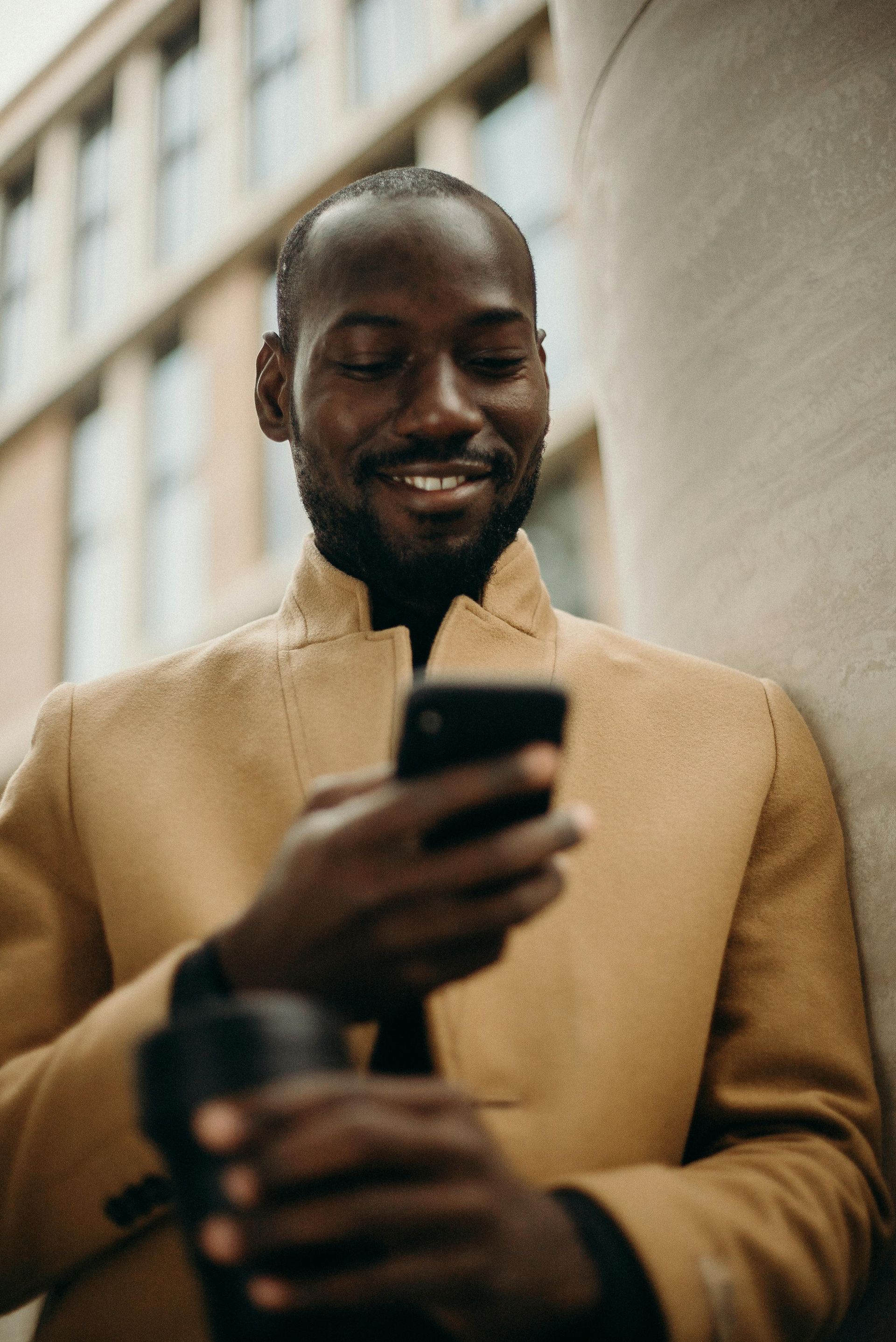 Man in tan jacket smiles while looking at a phone. Leans against a concrete column with a building in the background. Holds a coffee mug.