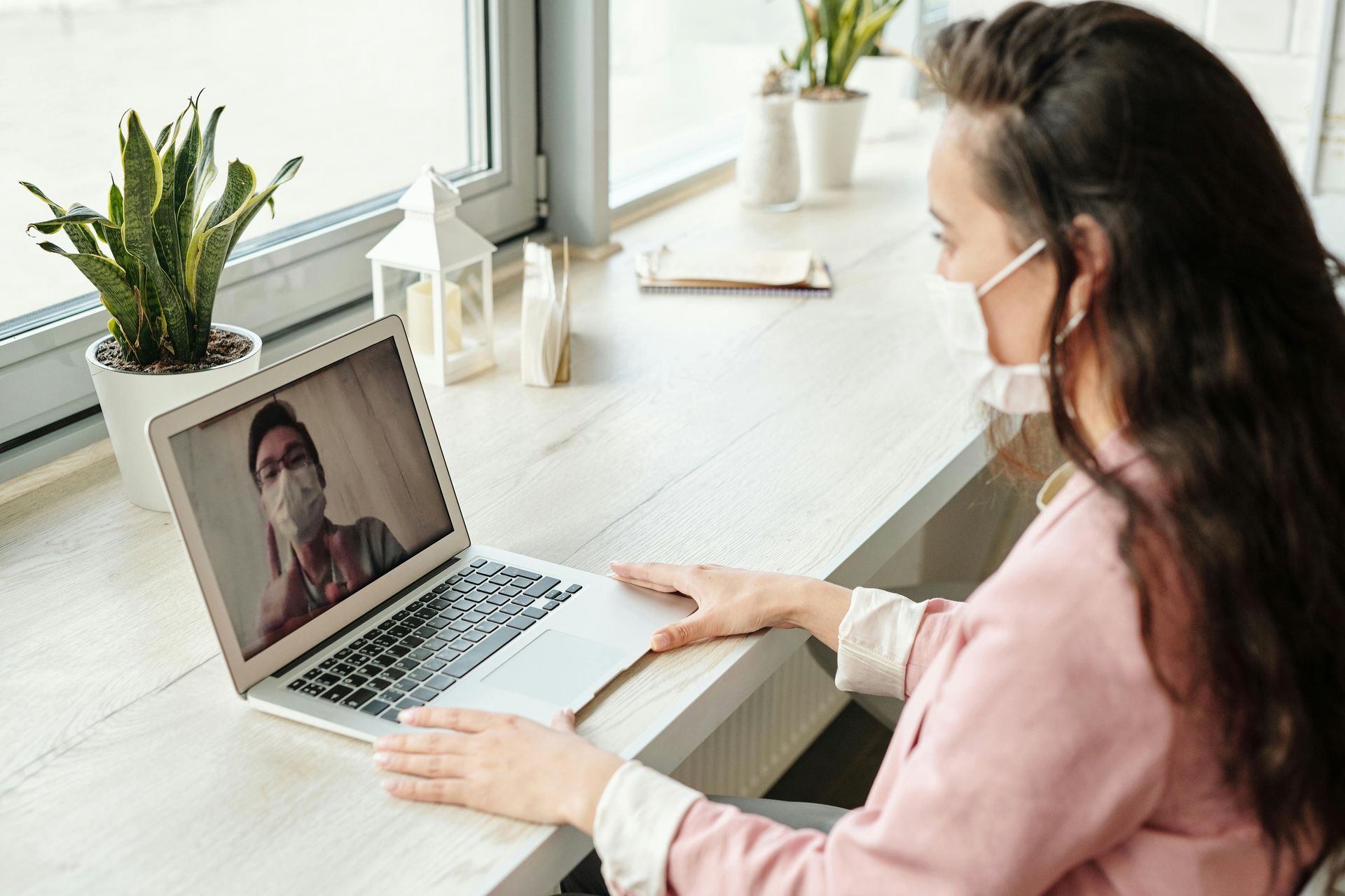 Woman in pink shirt and face mask video chatting on laptop by window.