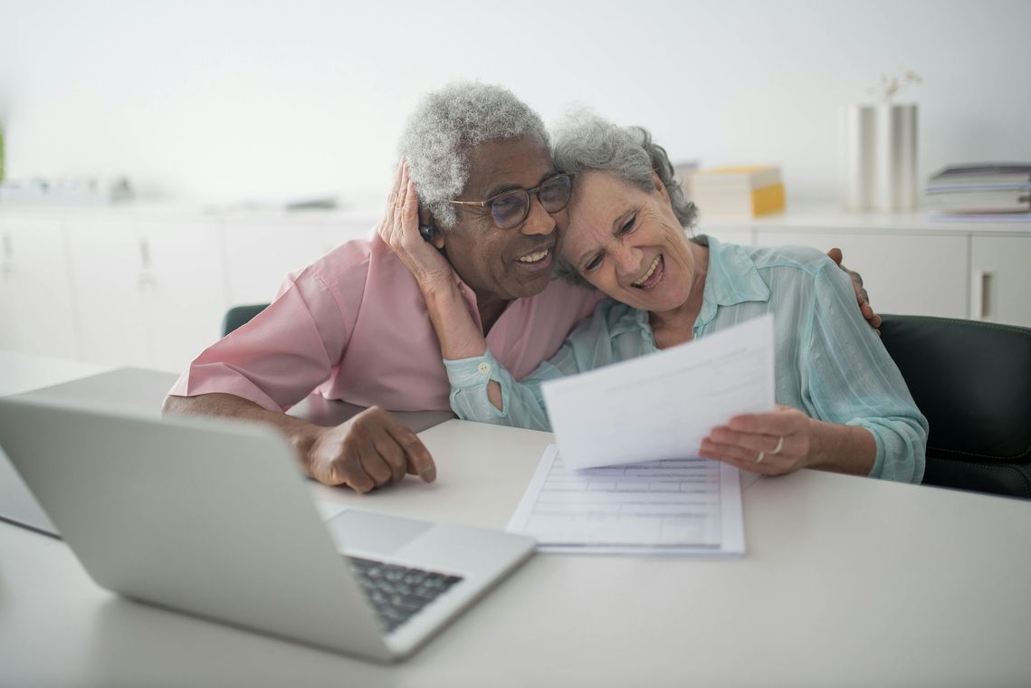 Smiling couple looking at paperwork, one with arm around the other at a table with a laptop.