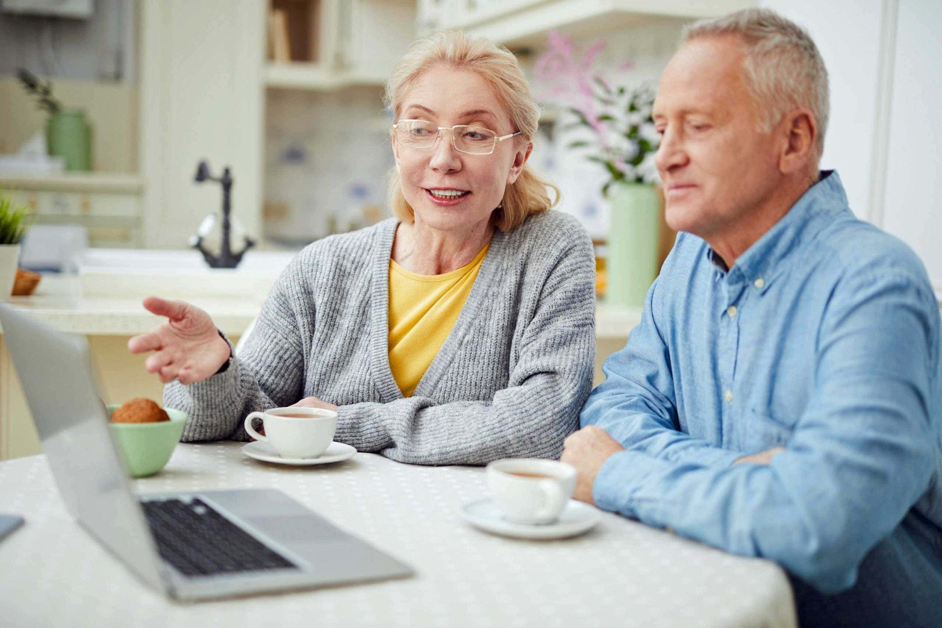 An elderly couple is sitting at a table looking at a laptop.