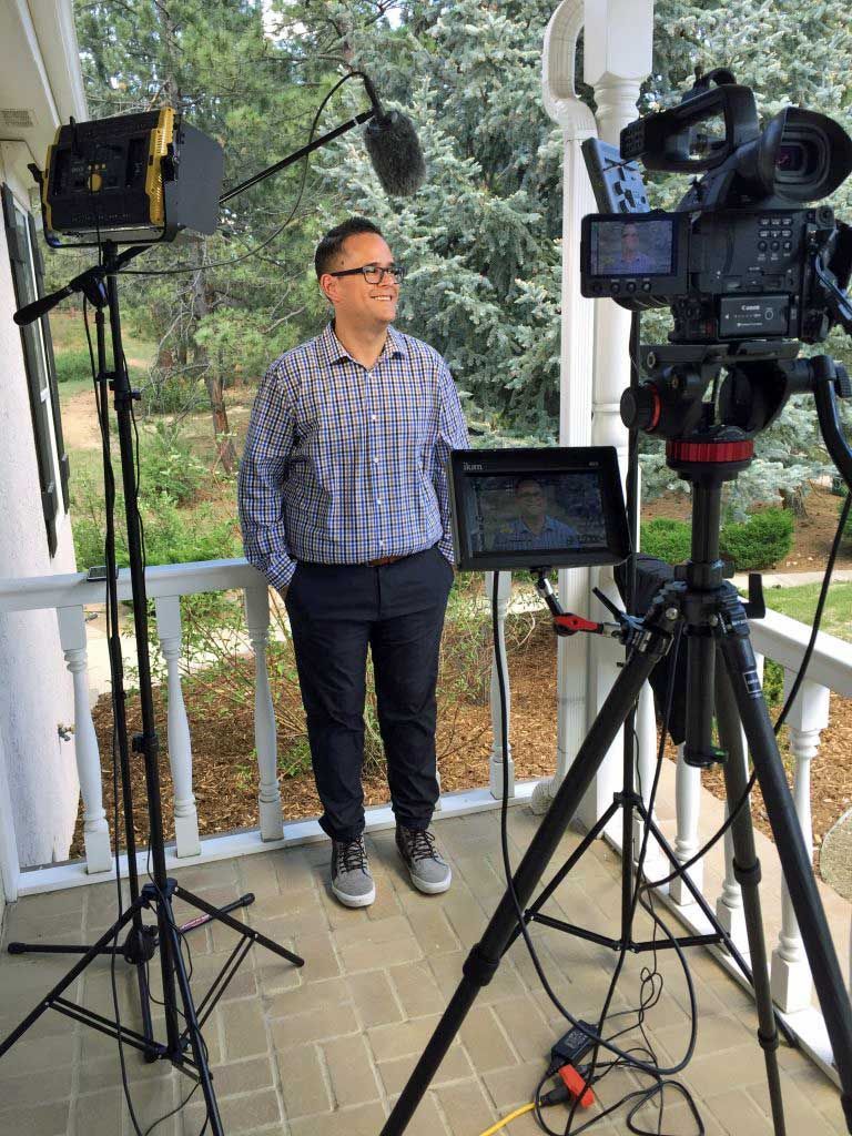 A man is standing on a porch in front of a camera.