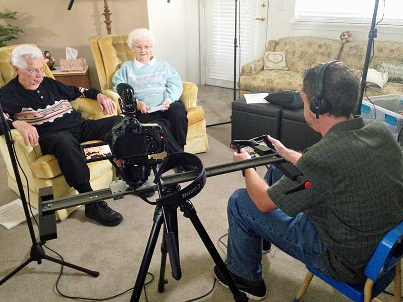 A man is sitting in front of a camera talking to two elderly people