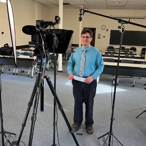 A man in a blue shirt and tie is standing in front of a camera