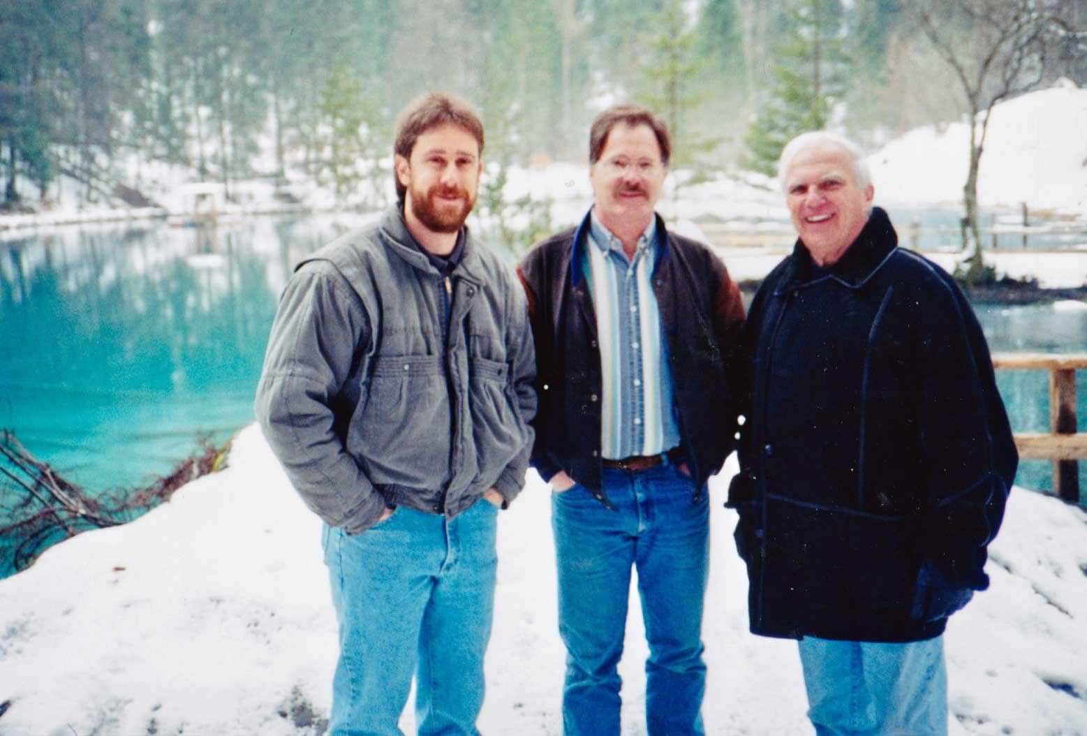 Three men are standing in the snow in front of a lake.