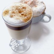 A glass of cappuccino next to a cup of cappuccino on a table.