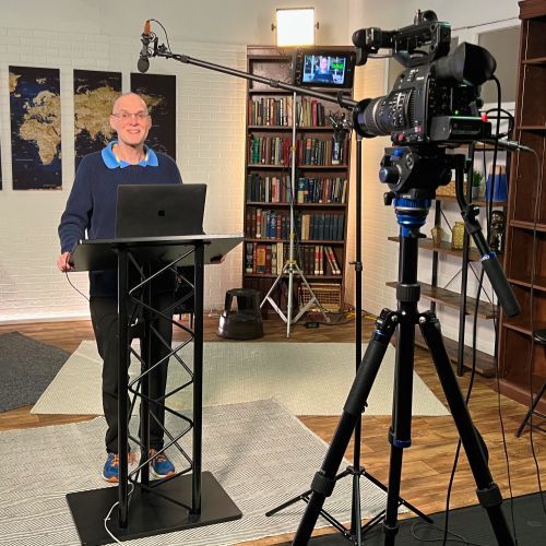 A man is standing at a podium with a laptop in front of a camera.