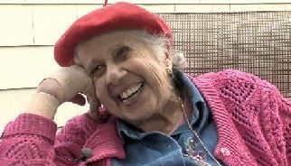 An elderly woman wearing a red hat and a pink sweater is smiling.