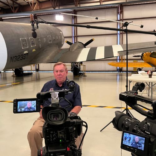 A man is sitting in front of a camera with a plane in the background.