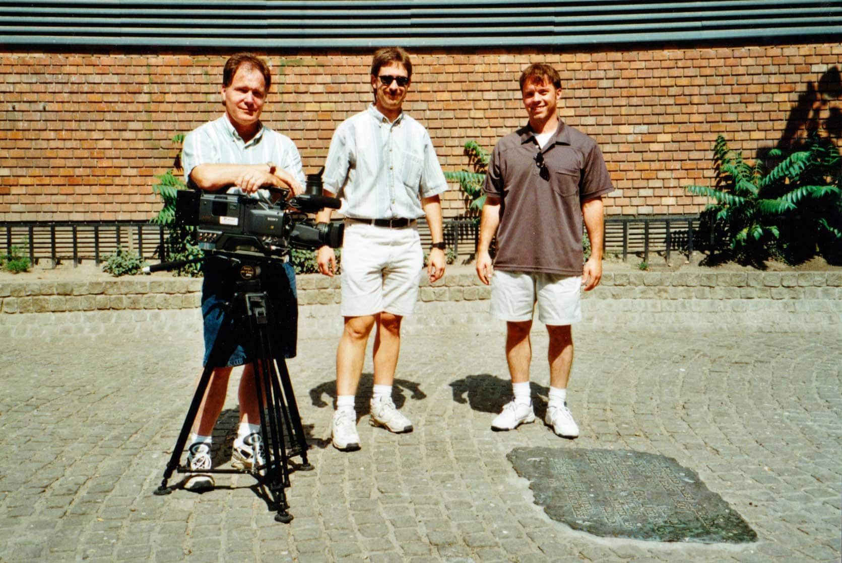 Three men standing in front of a camera on a tripod