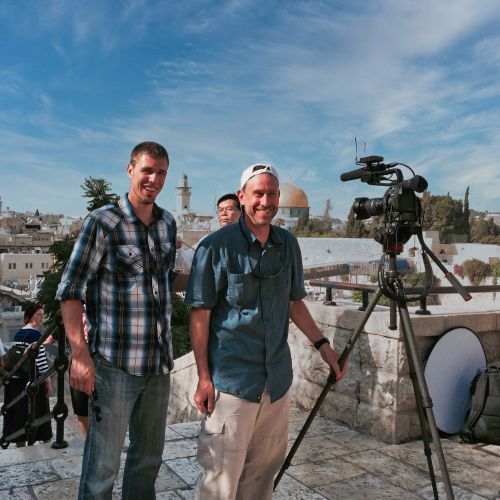 Two men standing in front of a camera on a tripod