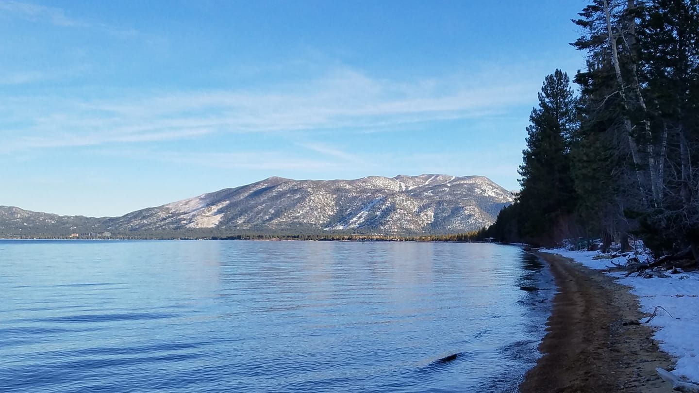 Snowy mountain lake with calm blue water, pine-covered shoreline, and a clear sky