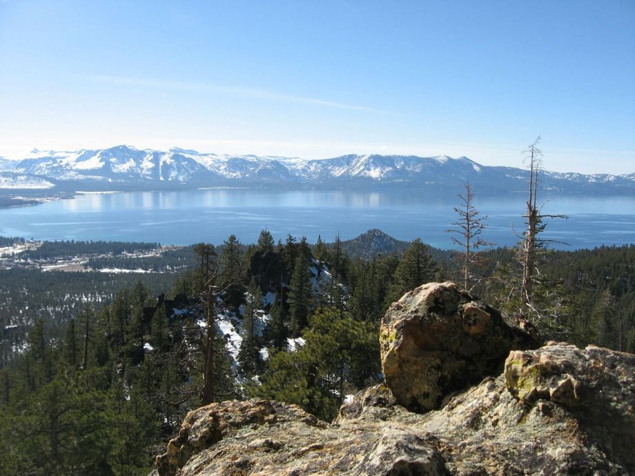 Snowy mountain range across a blue lake, with pine forest and rocky foreground under a clear sky.
