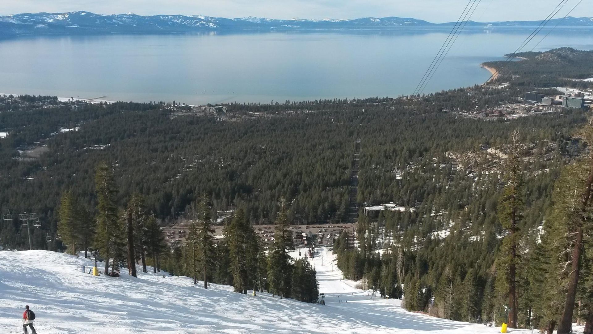 Snowy ski slope overlooking a forested hillside and a blue lake under a clear sky.
