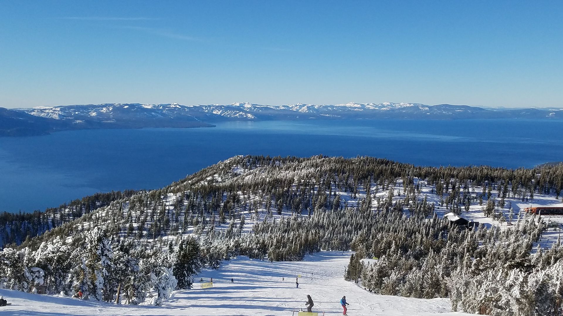 Snowy mountain ridge overlooking a blue lake with distant snow-capped hills under a clear sky.