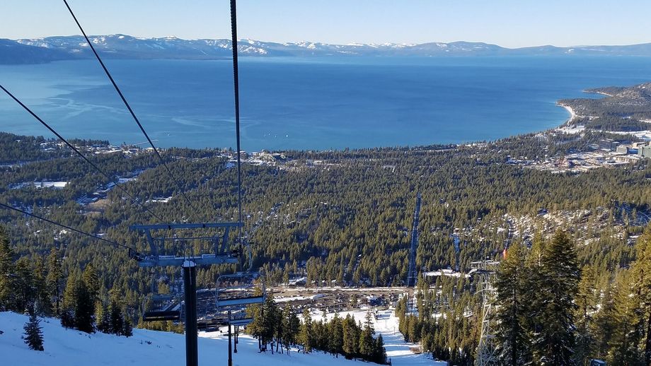 Snowy mountain overlook above a forested lakeside town with a blue lake and distant hills.