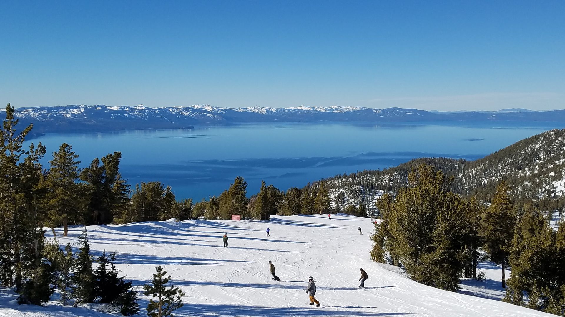 Snowy ski slope overlooking a blue lake and distant mountains under a clear sky