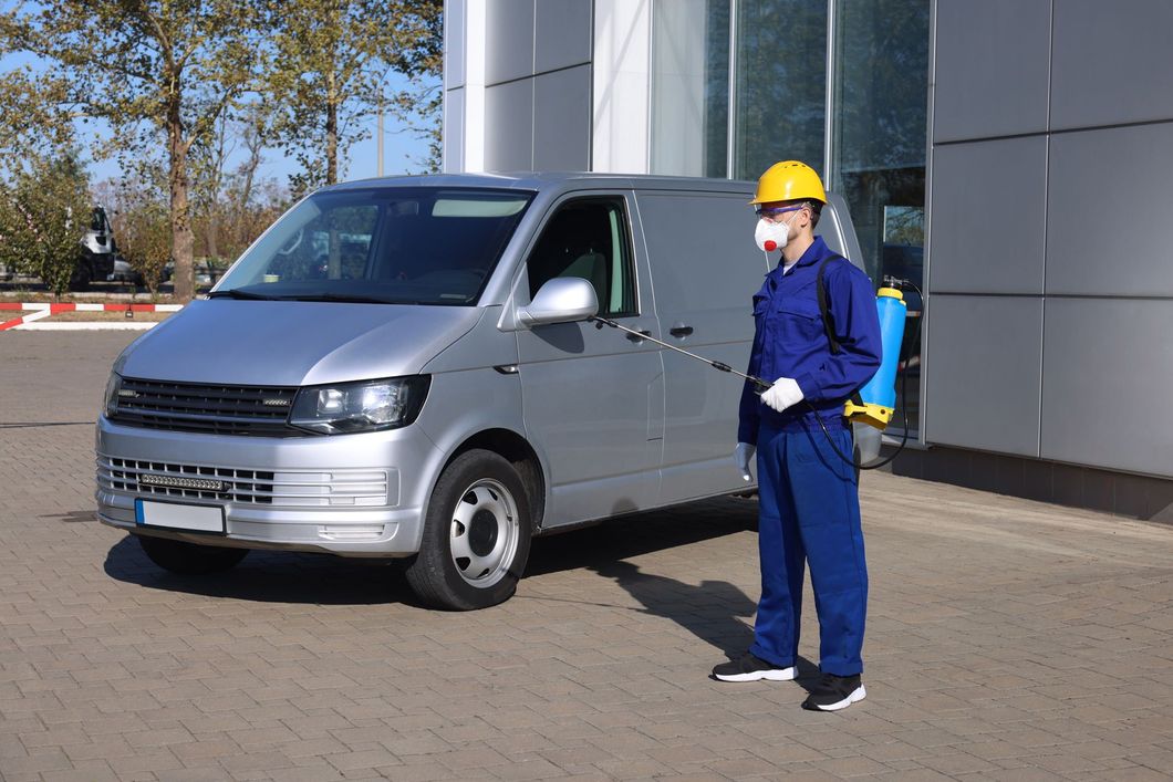 Worker in blue coveralls and yellow hard hat standing beside a silver van outdoors.