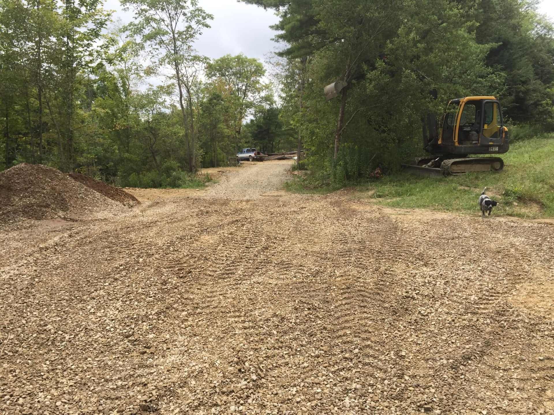 Cleared dirt road with wood chips, trees on both sides, and an excavator parked to the right.