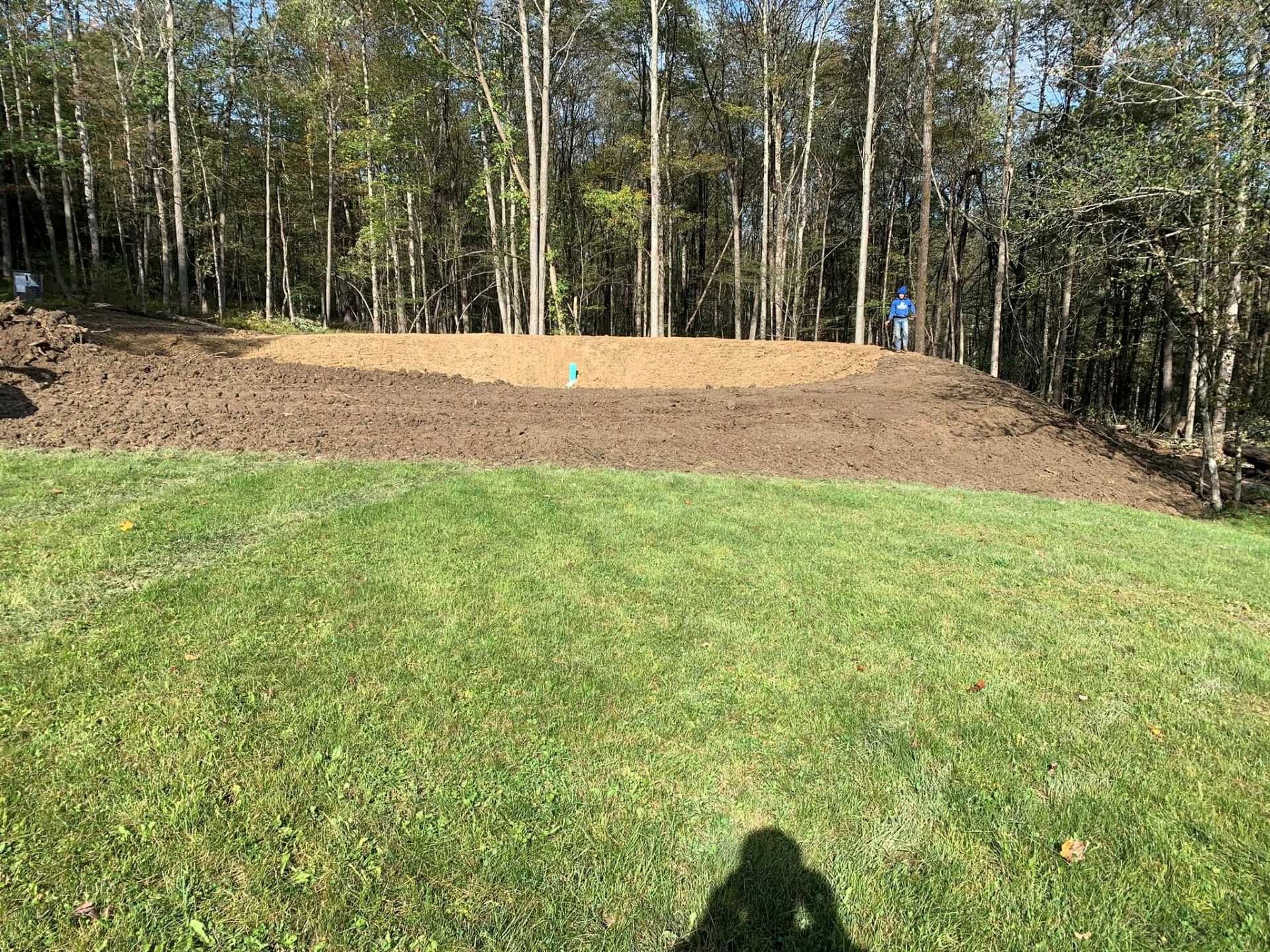 Grassy yard with a freshly graded dirt slope and trees in the background, two people standing near the top