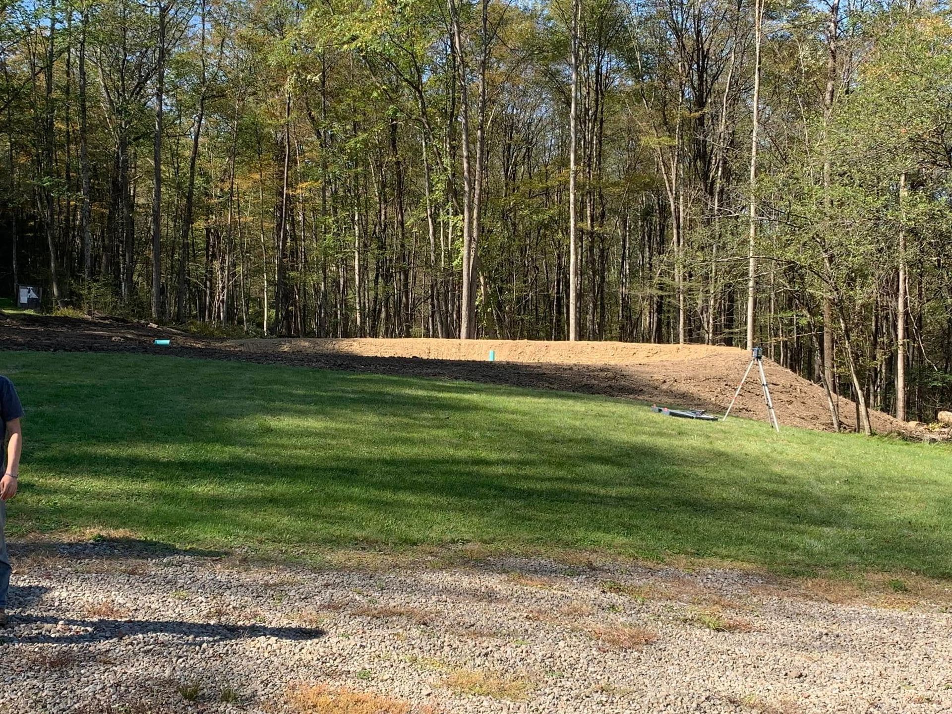 Grassy yard bordered by trees with a gravel drive in the foreground and a partial person at left edge