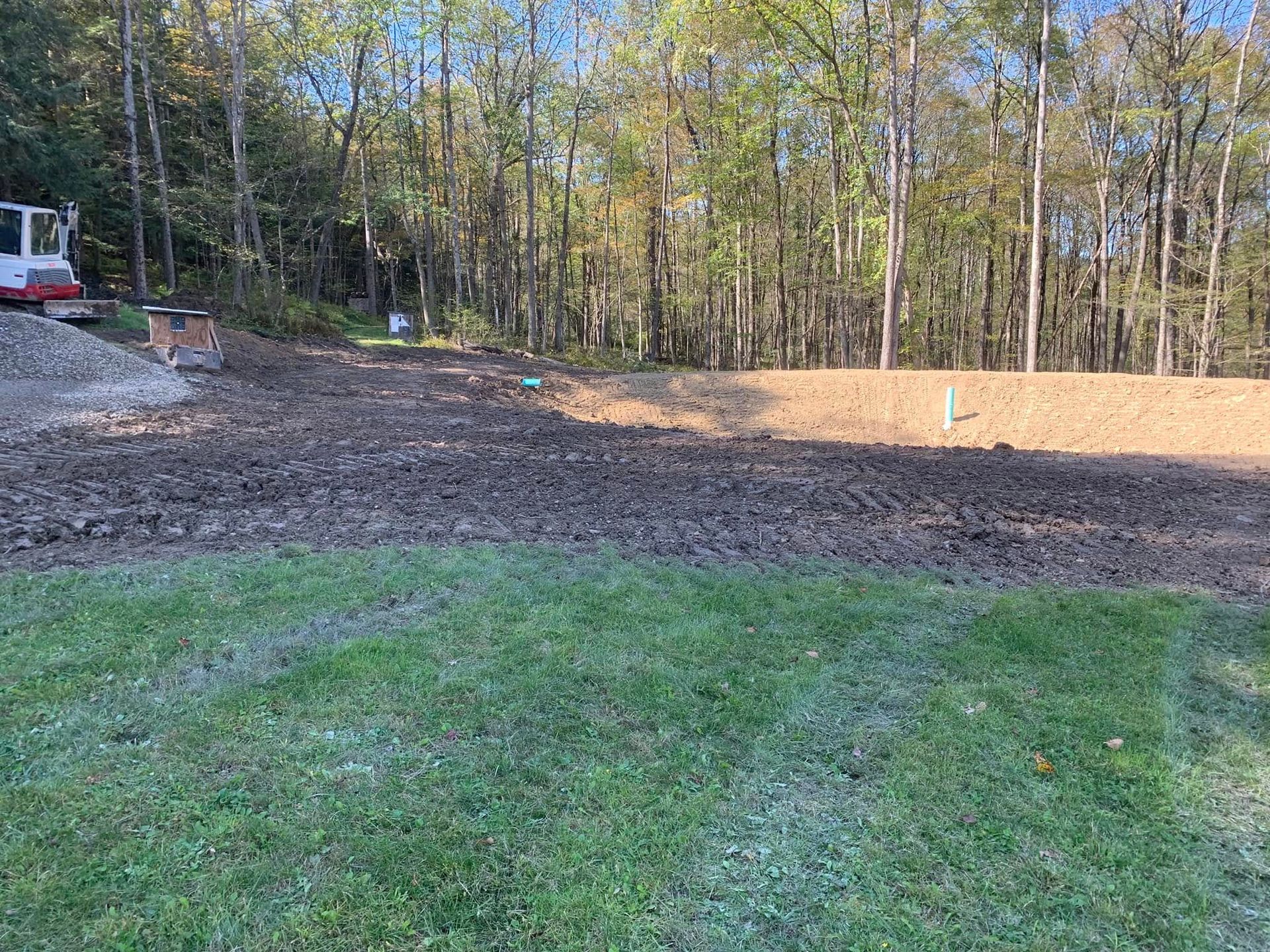 Cleared dirt lot beside a grassy yard and wooded edge, with a white truck at left and fresh soil berm in back