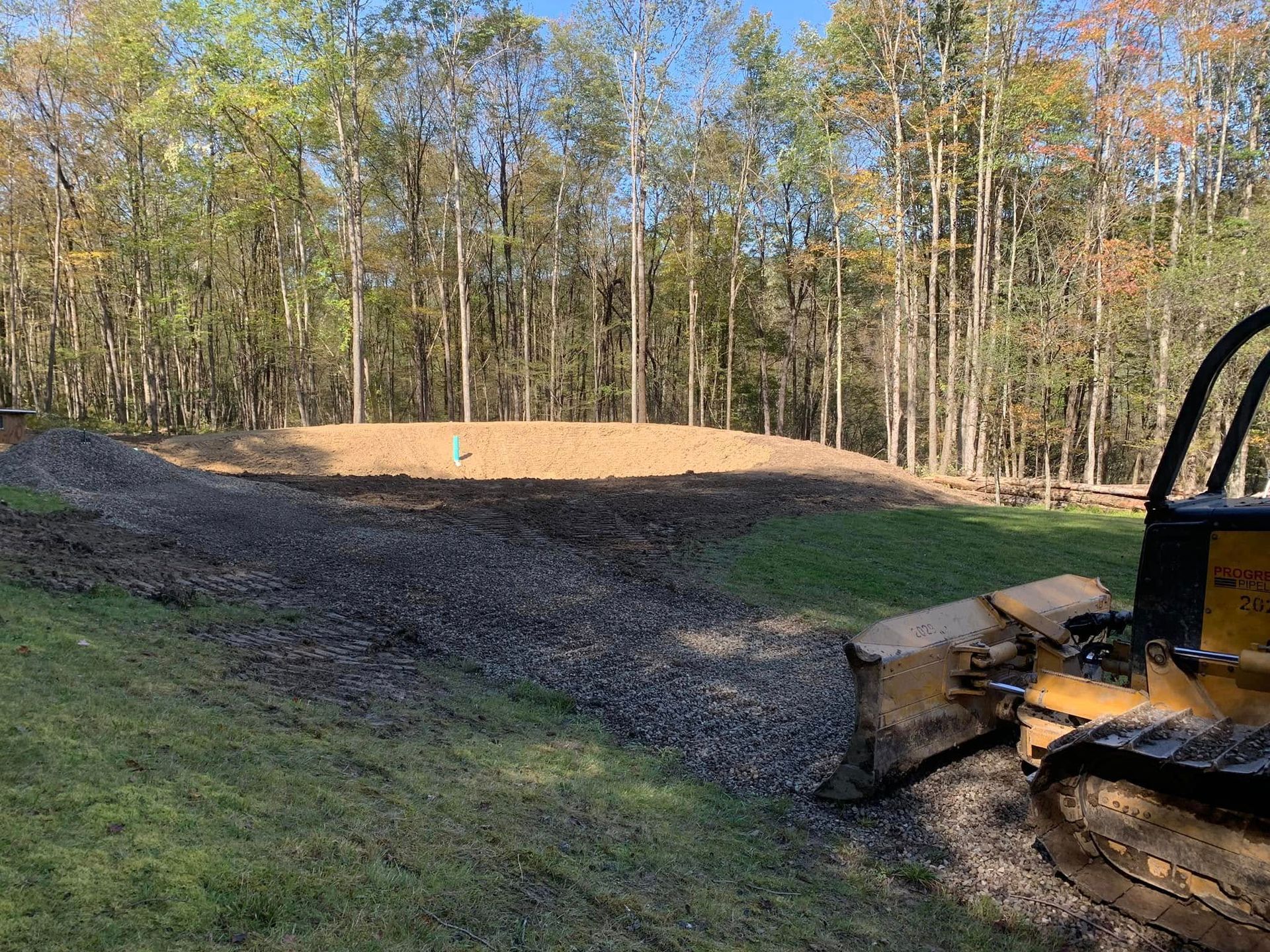 Bulldozer grading a dirt mound beside a wooded yard on a sunny day