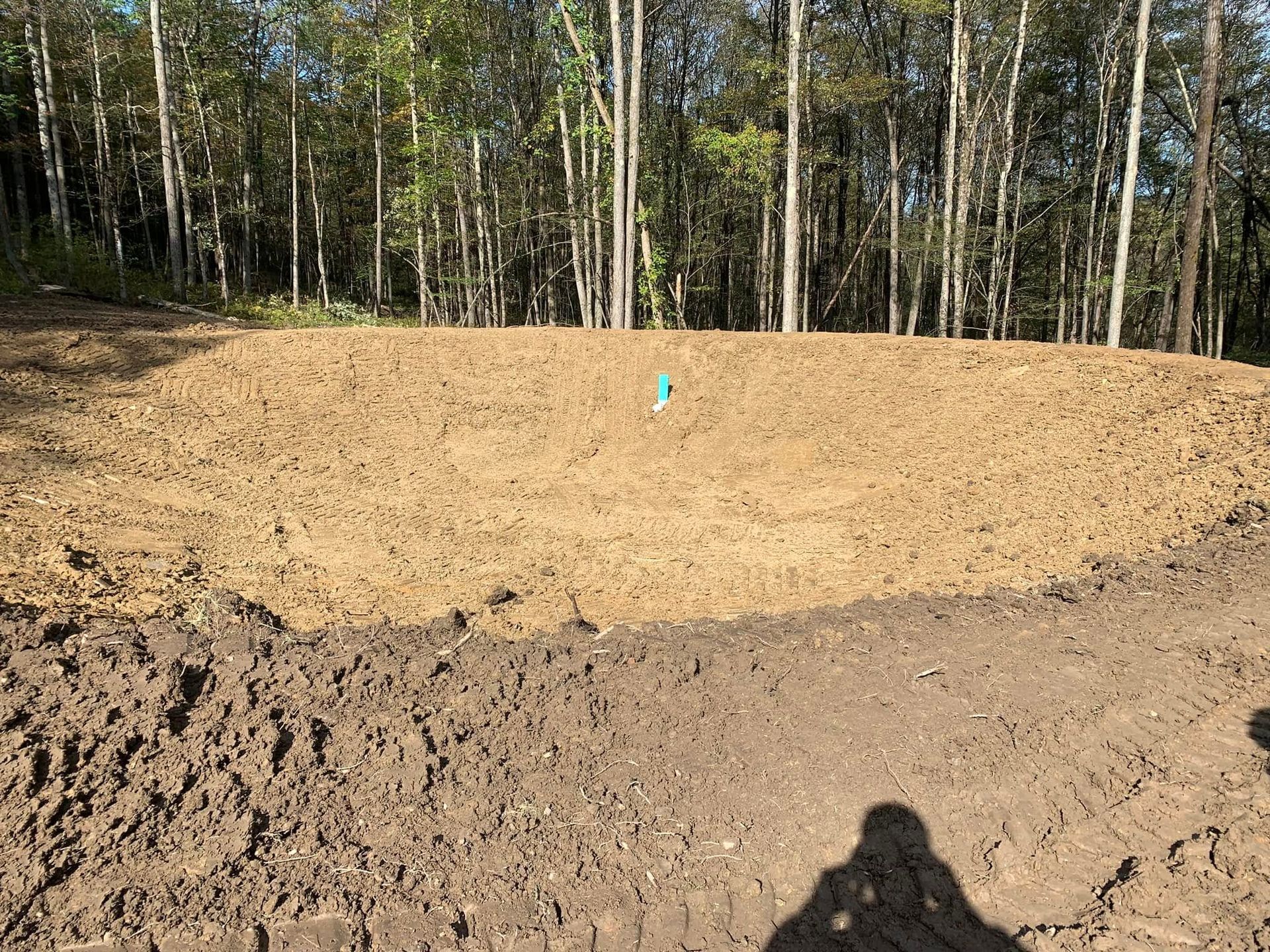 Freshly graded dirt mound in a wooded area with a small blue marker in the center.
