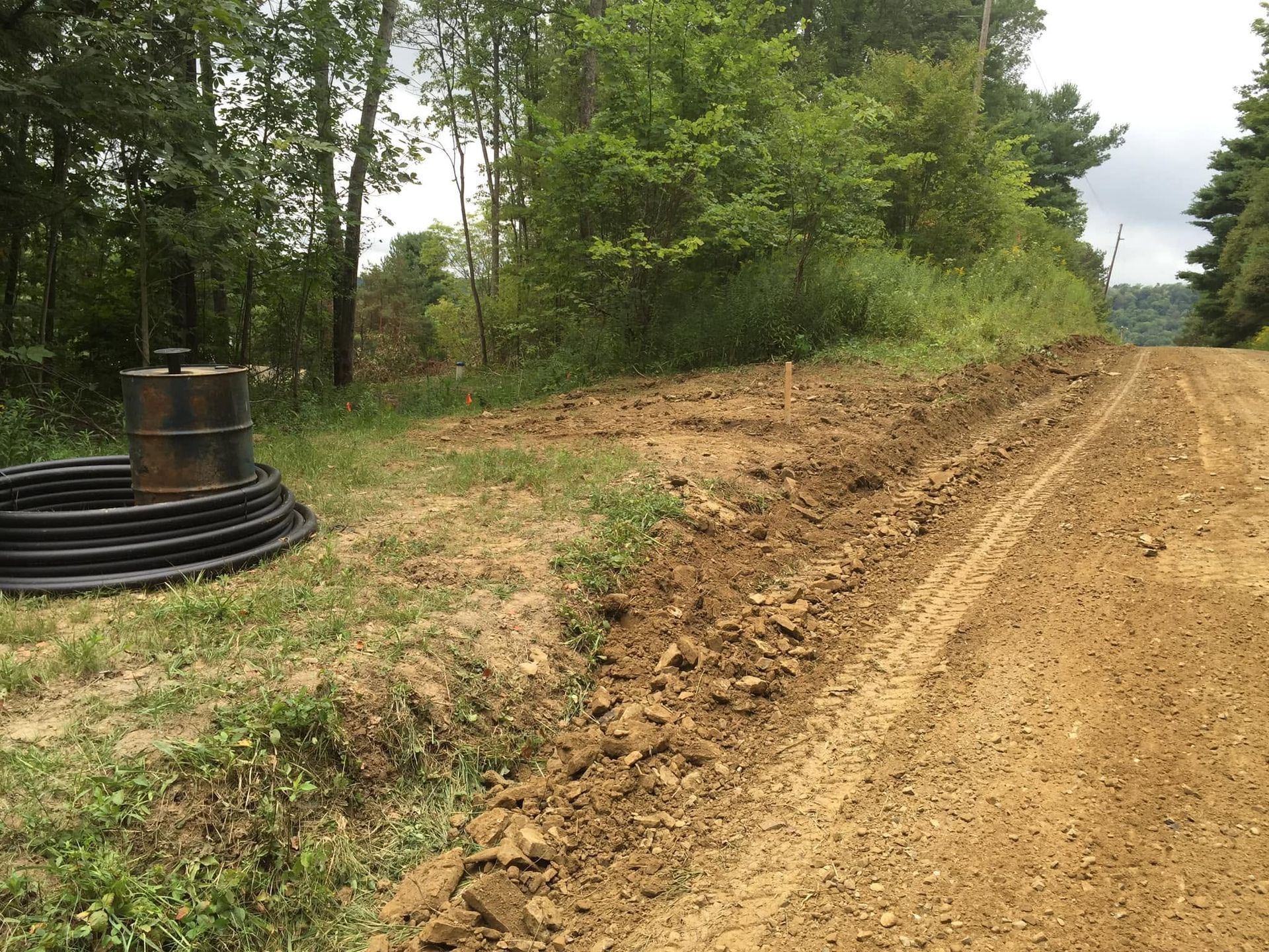 Dirt road under construction beside a wooded area, with coiled black pipe on the left.