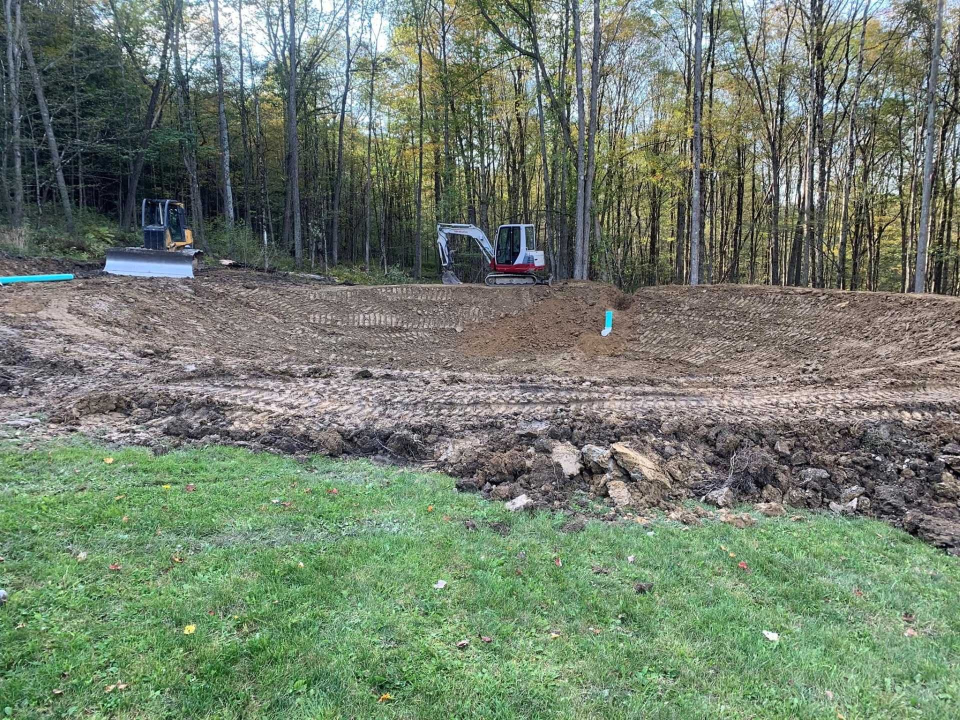 Cleared dirt lot with a small excavator and trees in the background near a grassy foreground