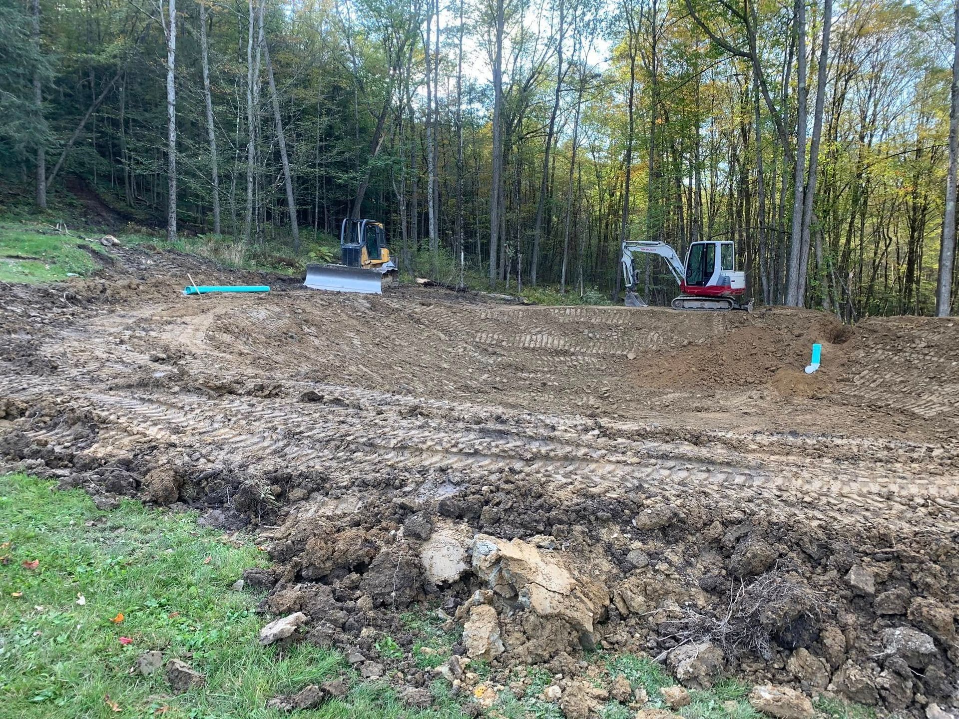 Cleared dirt lot with muddy tracks, a small excavator, and trees in the background