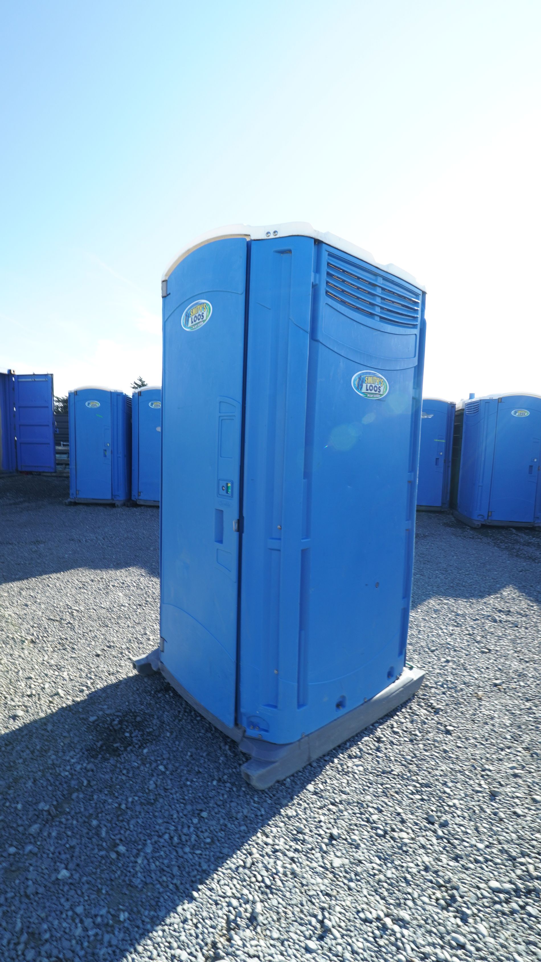 Blue portable toilet on gravel with others in the background under a clear sky.